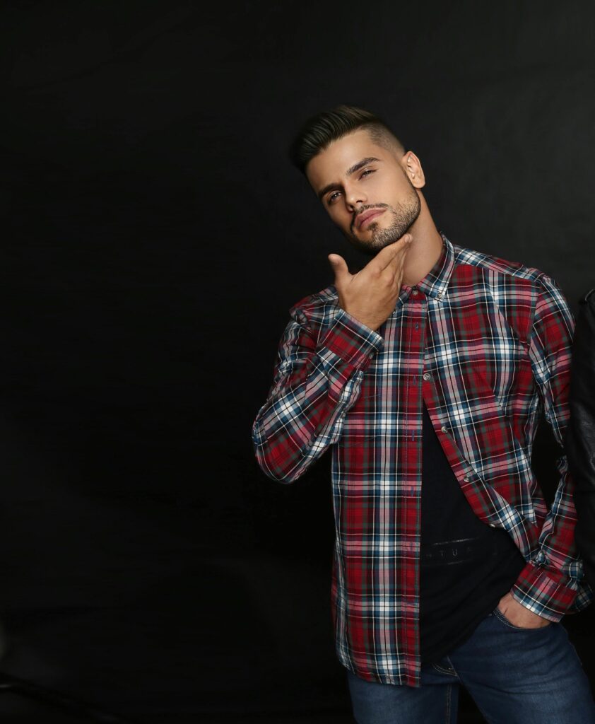 Stylish young man posing in a checked flannel shirt against a black background in a studio setting.